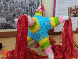 A colorful star-shaped piñata with red streamers is displayed in front of a black and white photograph of various piñatas.
