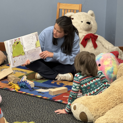 A child sitting on the floor among large stuffed animals, being read a book. 