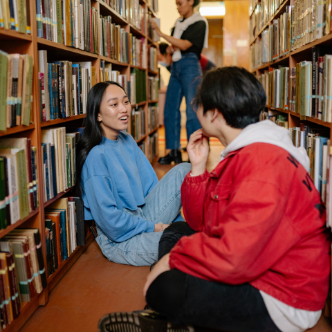 Two teens sitting on the floor, chatting in-between library stacks, while another teen is browsing in the background. 
