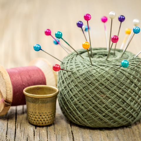 A thimble sits next to a ball of green yarn with colorful pins stuck in it, and a spool of pink thread is in the background.
