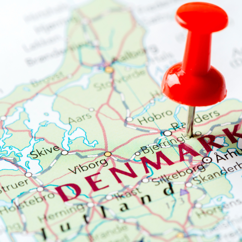 A red thumbtack is placed on a map of Denmark, marking the country." DENMARK" is prominently displayed on the map.