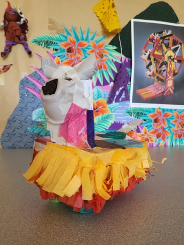 A handmade piñata shaped like a white animal's head with colorful tissue paper fringe rests on a table in front of a vibrant tropical-themed backdrop.
