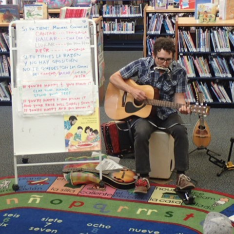 A person plays an acoustic guitar while sitting on a colorful rug in front of a whiteboard with writing and a bookshelf filled with books.