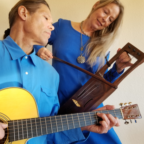 Musician Scott Hongell-Darsee plays an acoustic guitar while Johanna Hongell-Darsee holds a lyre behind them.