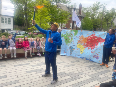 A person holding a lit torch and a world map with children sitting on a bench in front of them.
