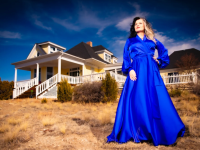 Musician Wendy Rule in a flowing blue satin robe stands on a dry, grassy hill in front of a large yellow house with a white porch and dark roof under a bright blue sky with scattered clouds.