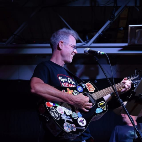 A person plays an electric guitar on a stage with dark lighting.