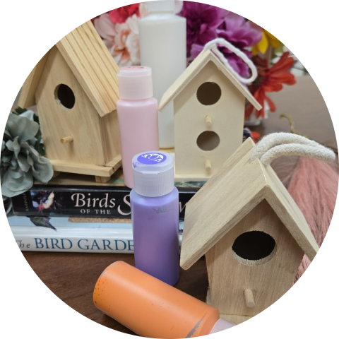 Three unfinished wooden birdhouses with bottles of craft paint in pink, purple, and orange, arranged on a stack of books with a floral arrangement in the background.