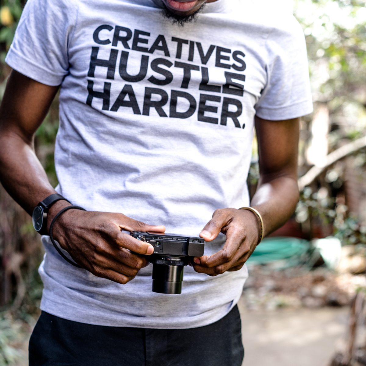 man holding a camera and wearing a shirt that says "Creatives Hustle Harder"